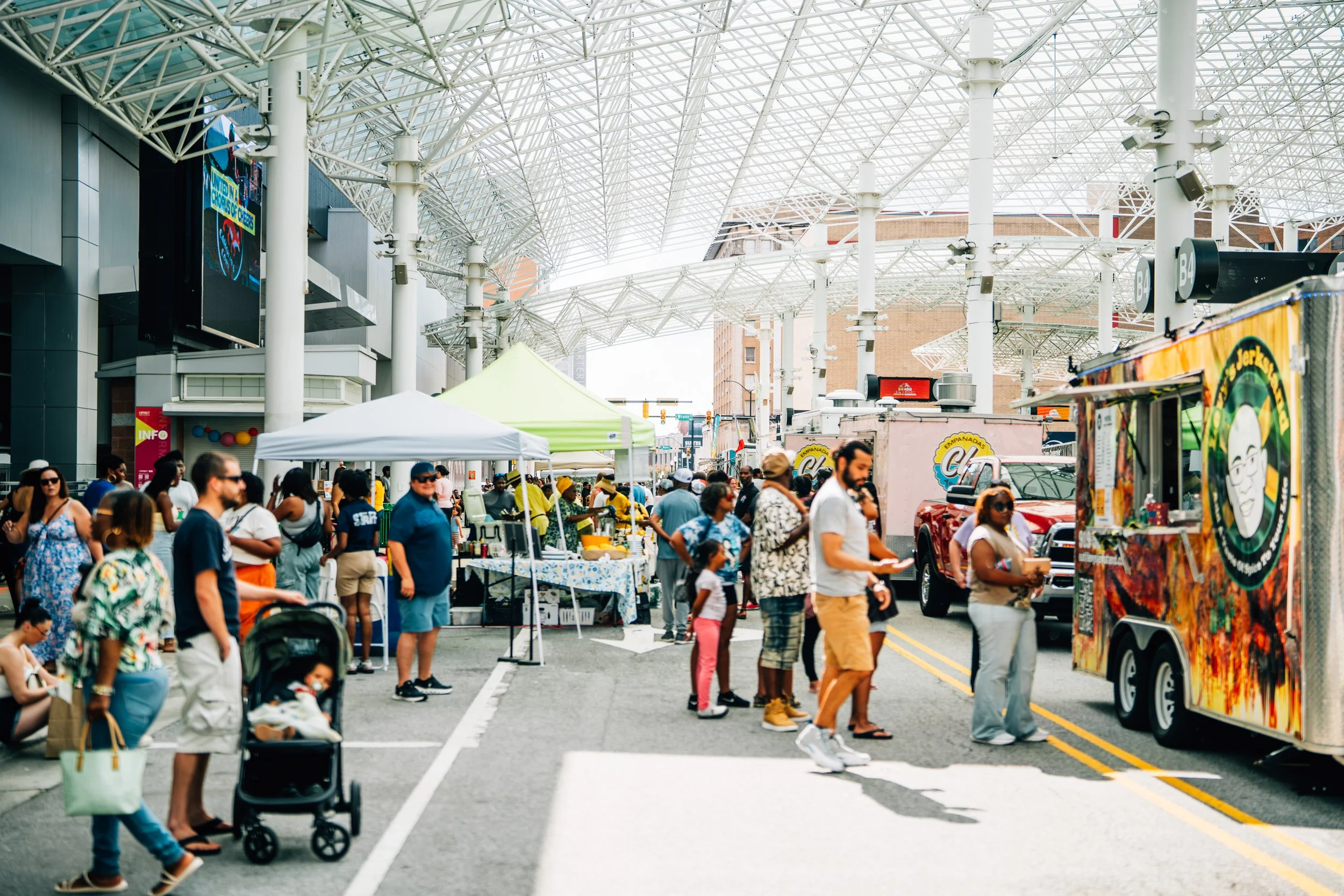 A busy outdoor street market with people shopping under a large modern, white metal lattice roof. There are food and vendor stalls, some with tents, and a food truck on the street. People are walking, talking, and browsing, with a diverse crowd and clear sky.