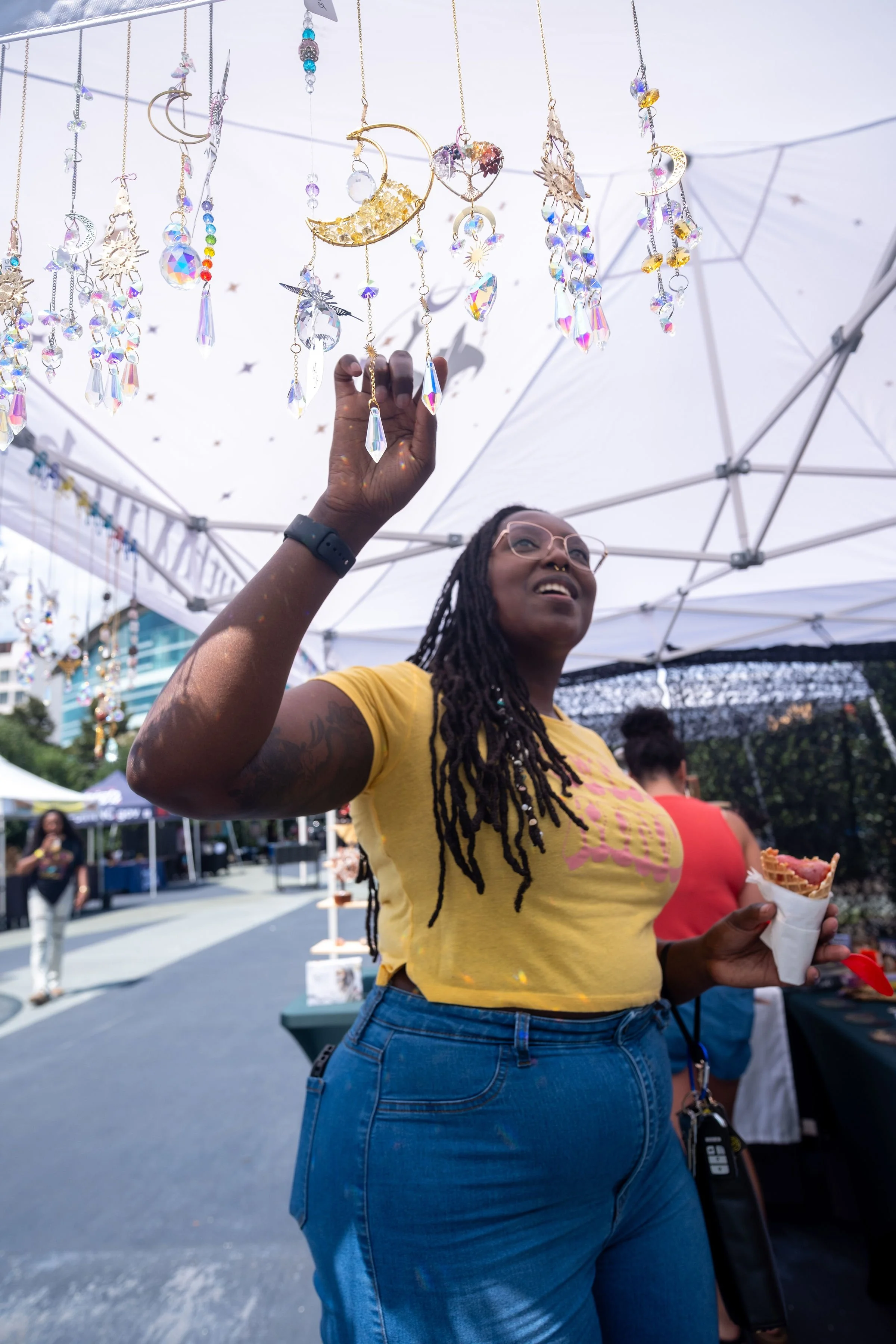 A woman with long dreadlocks wearing a yellow shirt and blue jeans is at an outdoor event, reaching up to browse hanging decorative glass ornaments and jewelry under a white canopy. She is holding a waffle cone with food.