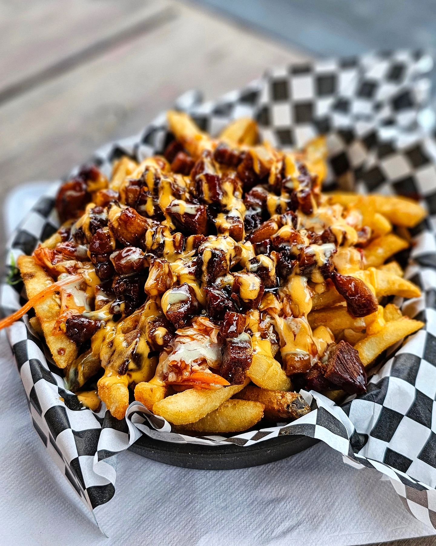 Basket of loaded French fries topped with cheese, barbecue sauce, and bacon bits, served on checkered paper lining.