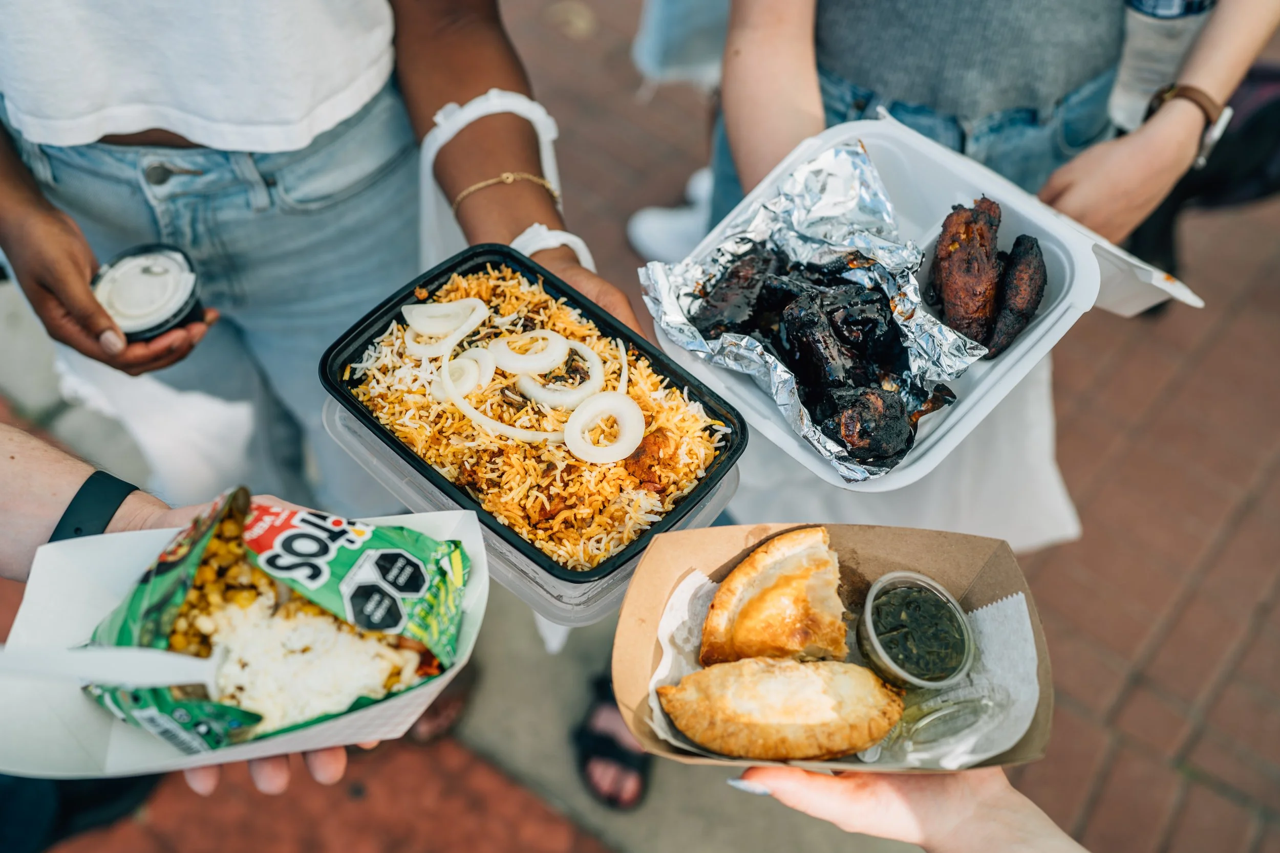 People holding takeout food containers with rice, grilled meats, rolls, and a drink.