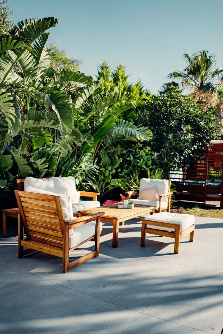 Outdoor patio with wooden chairs and cushions, surrounded by lush green plants, under a clear blue sky.