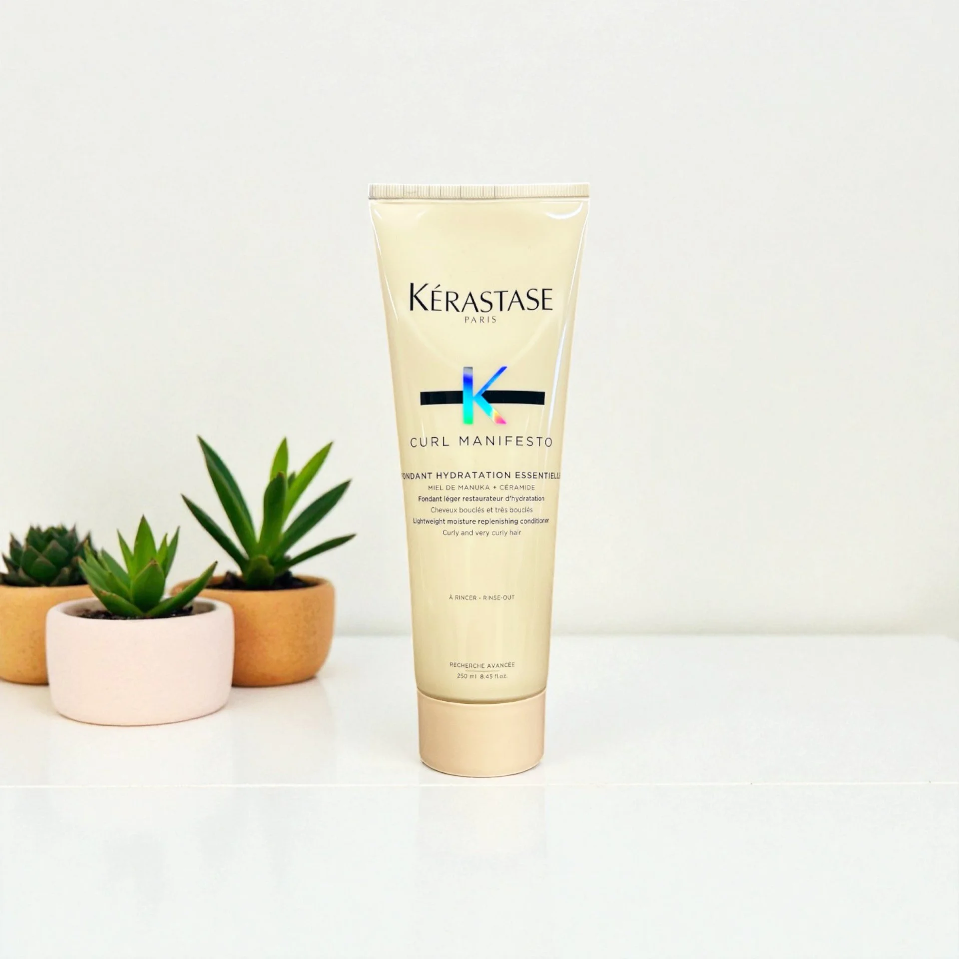 Kérastase Curl Manifesto lightweight moisture conditioner in a beige tube, placed on a white surface with three small potted succulents in the background.
