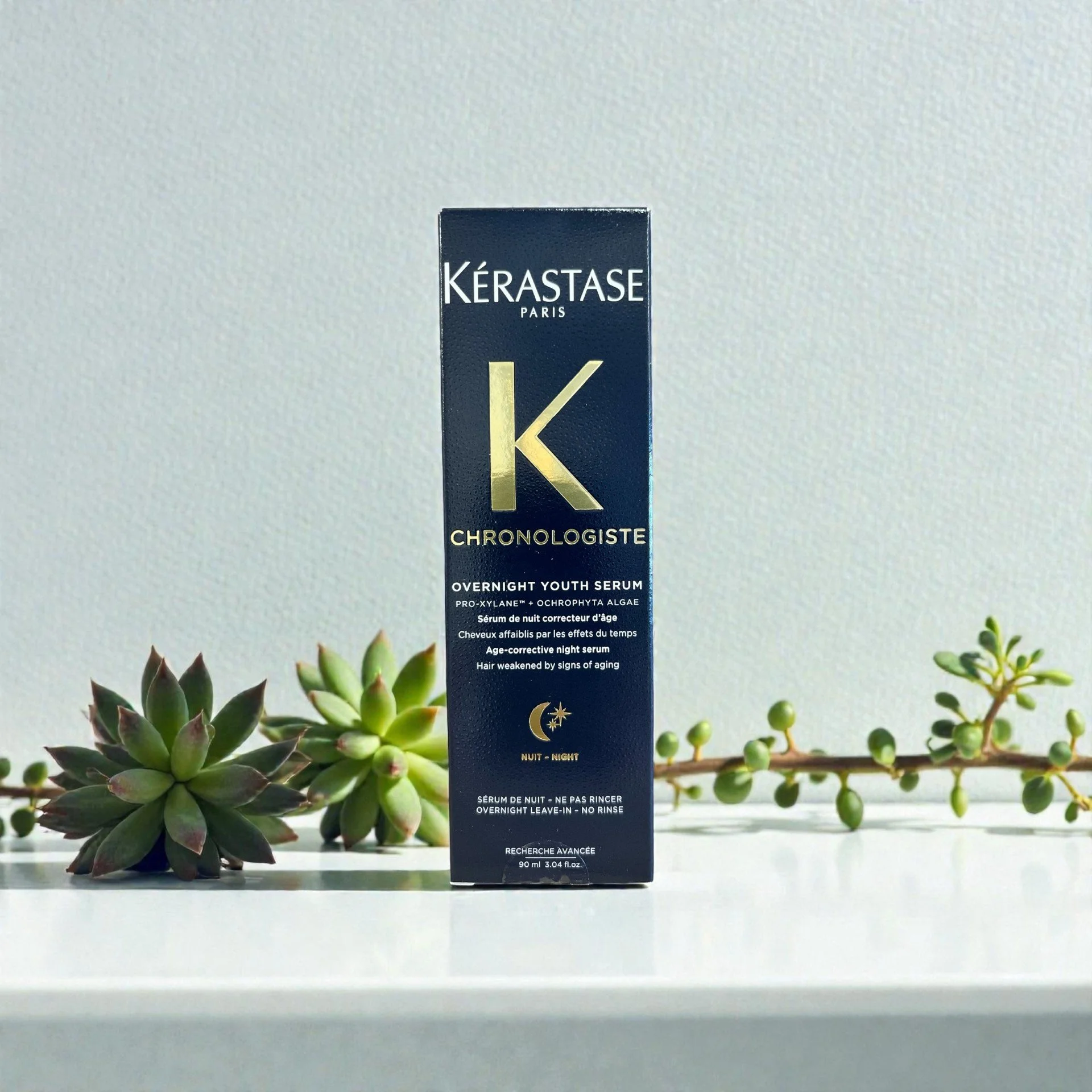 A black box of Kérastase Paris Chronologiste Overnight Youth Serum placed on a white surface with two succulent plants nearby, set against a plain white wall.