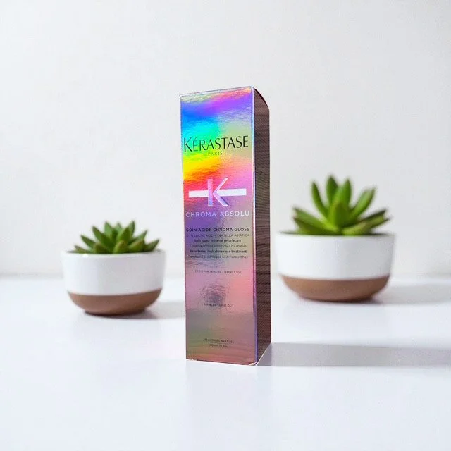 A colorful box of Kérastase Chroma Absolu gloss hair treatment positioned center with two potted succulents on either side against a plain white background.