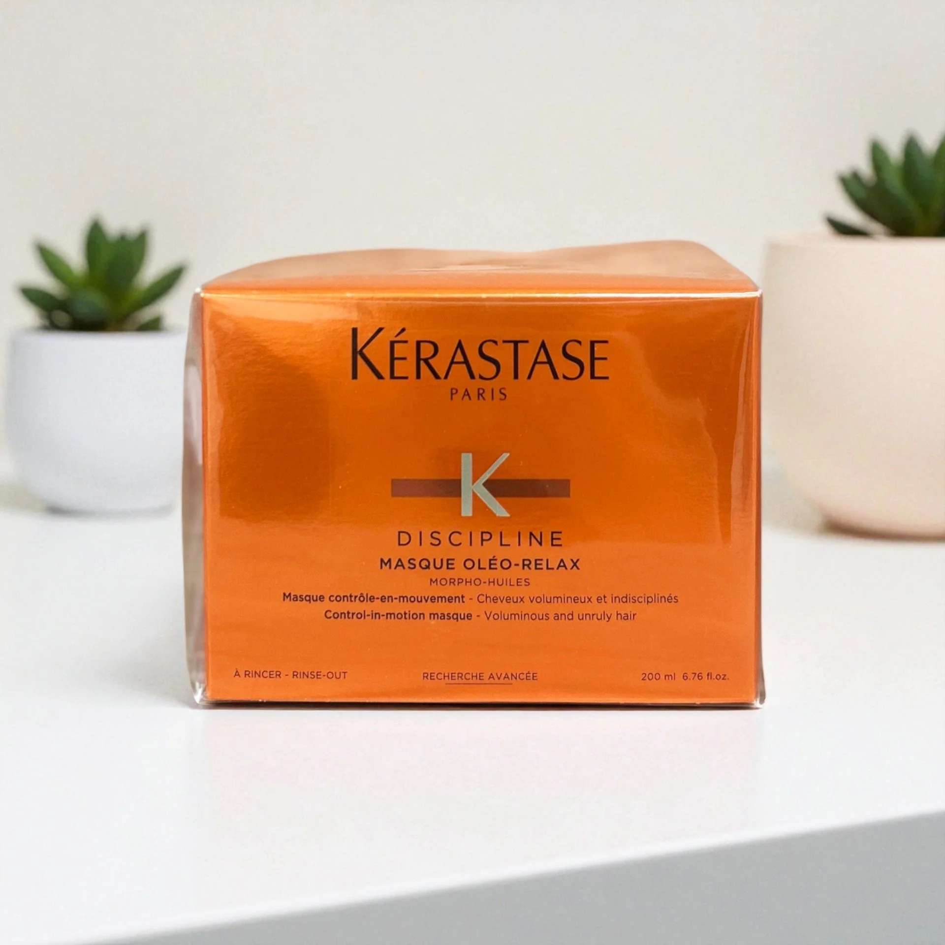 Orange box of Kérastase Paris Discipline Masque Oléo-Relax hair treatment on a white surface with two small potted succulents in the background.