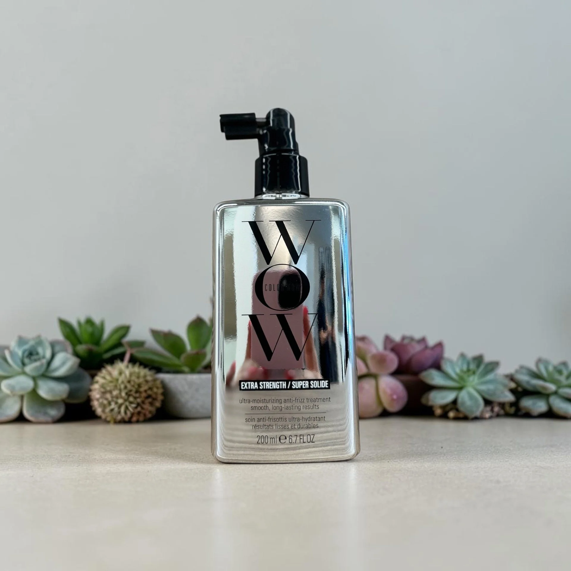 A bottle of Wowo Extra Strength Super Solid anti-frizz treatment standing on a white surface with a background of assorted green and purple succulents.