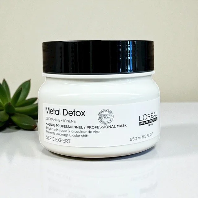 A white jar of L'Oréal Metal Detox professional mask with a black lid, placed on a white surface with a green plant in the background.