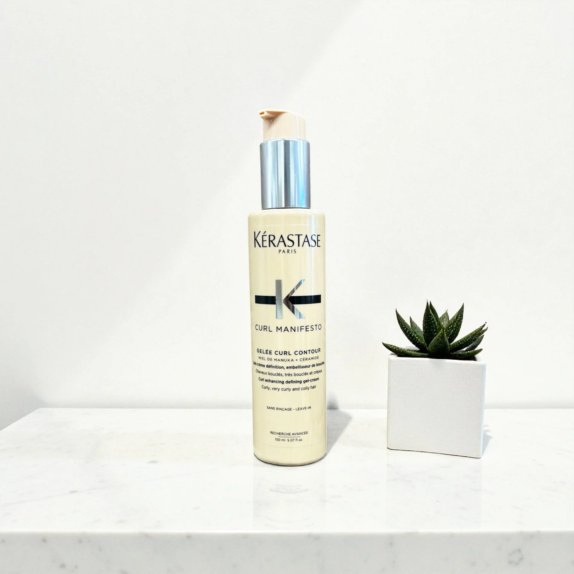 A bottle of Kérastase Paris Curl Manifesto gelée curl contour beside a small potted succulent plant on a white surface against a plain white background.