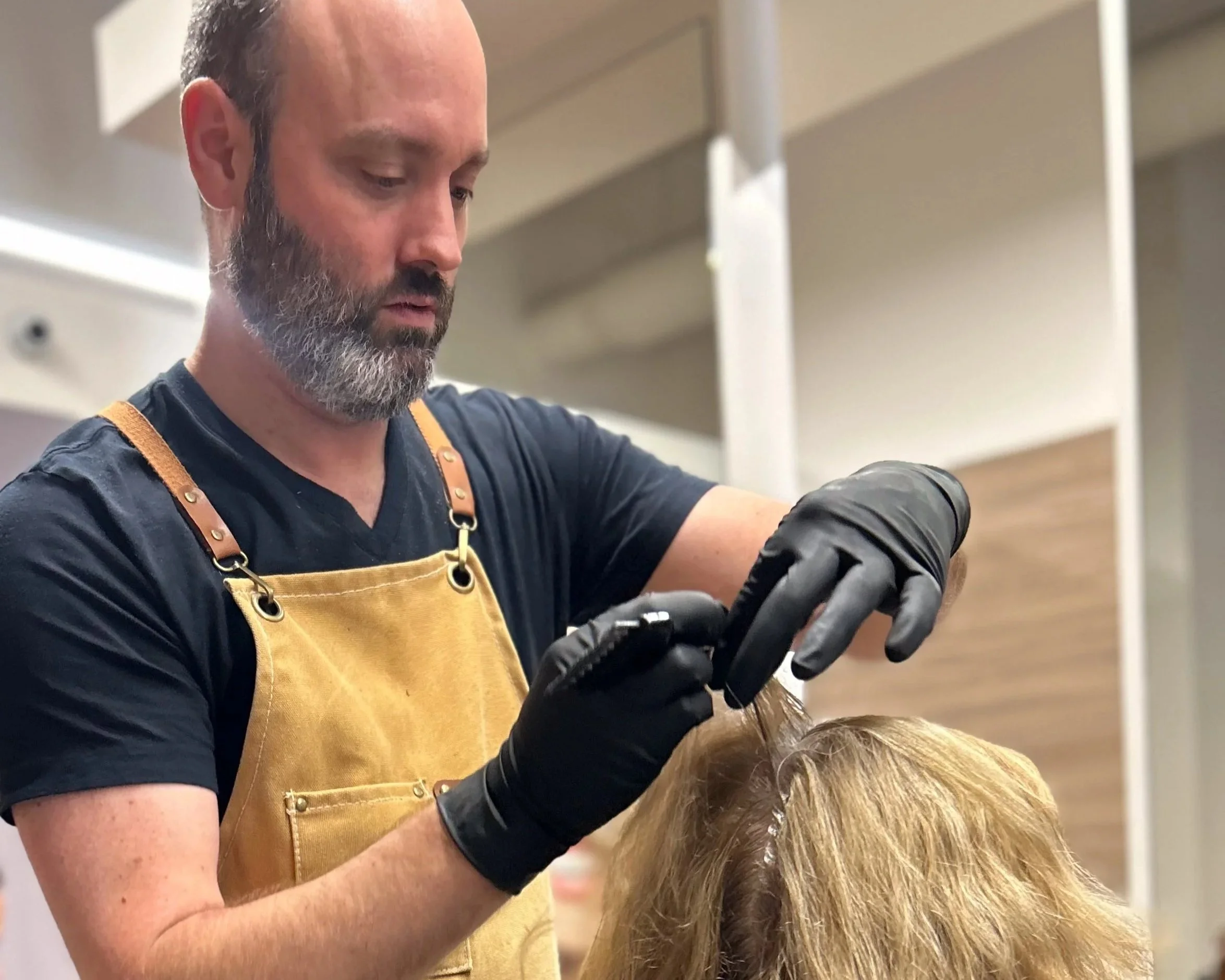 A stylist with a beard, wearing a black shirt, black gloves, and a tan apron, is applying highlights or coloring to a woman's blonde hair in a salon.