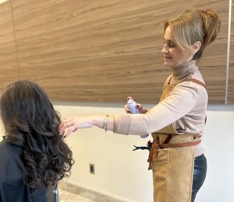 Woman in an apron and beige sweater applying makeup or face paint to another woman with long, curly dark hair in a room with wood paneling on the wall.