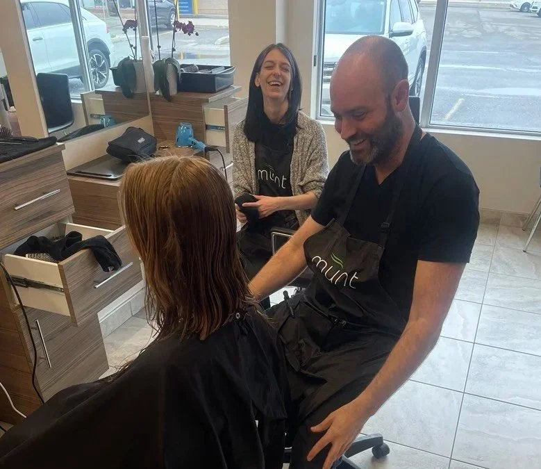 A barber and a woman in a salon, laughing and smiling while giving a haircut to a client with red hair, seated in a salon chair.