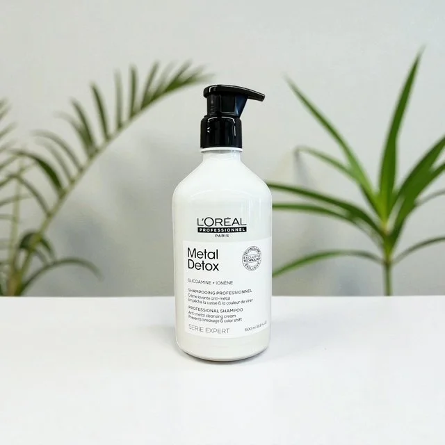 A white bottle of L'Oréal Professionnel Metal Detox shampoo with a black pump dispenser, placed on a white surface with green plants in the background.