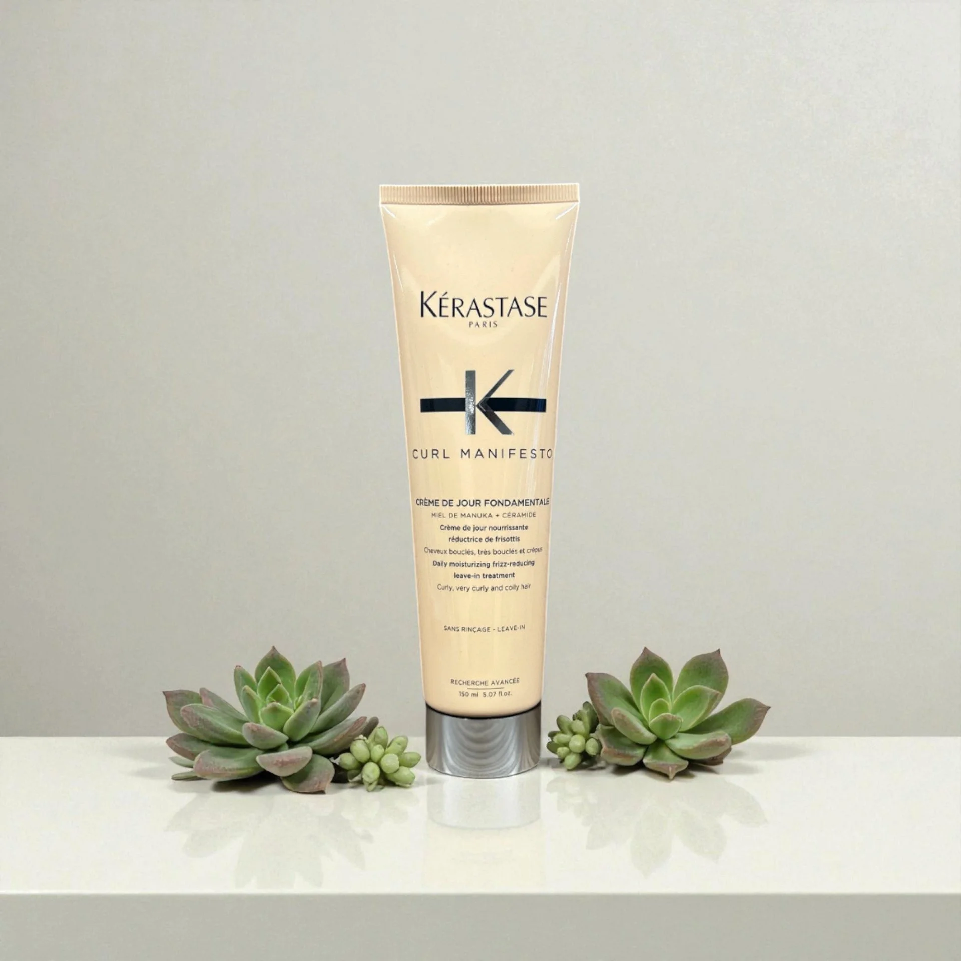 A tube of Kerastase Curl Manifesto leave-in conditioner placed between two small succulent plants on a white surface with a plain background.