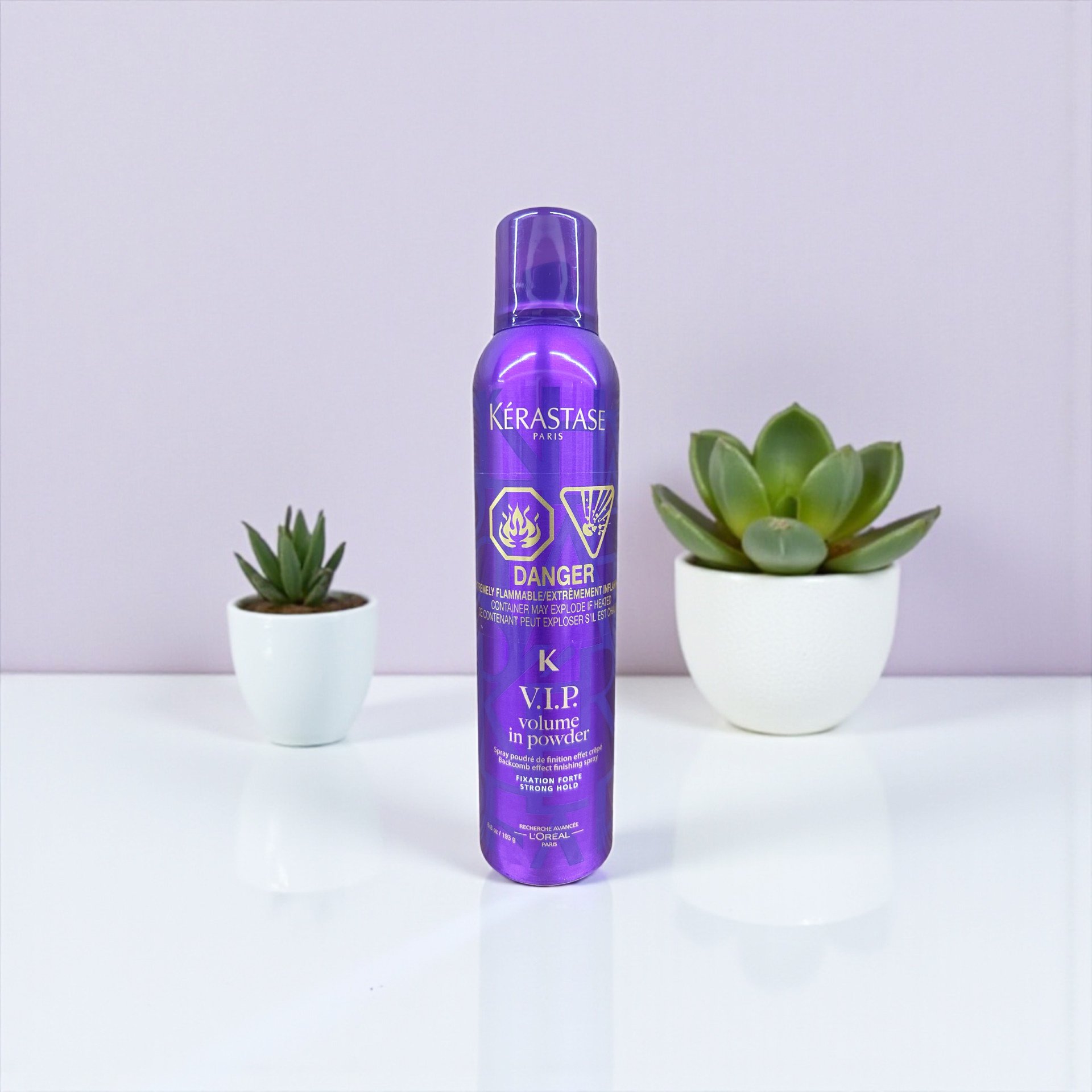 Purple can of Kérastase V.I.P. volume in powder spray with two potted succulents, one small and one larger, on white surface against plain white background.
