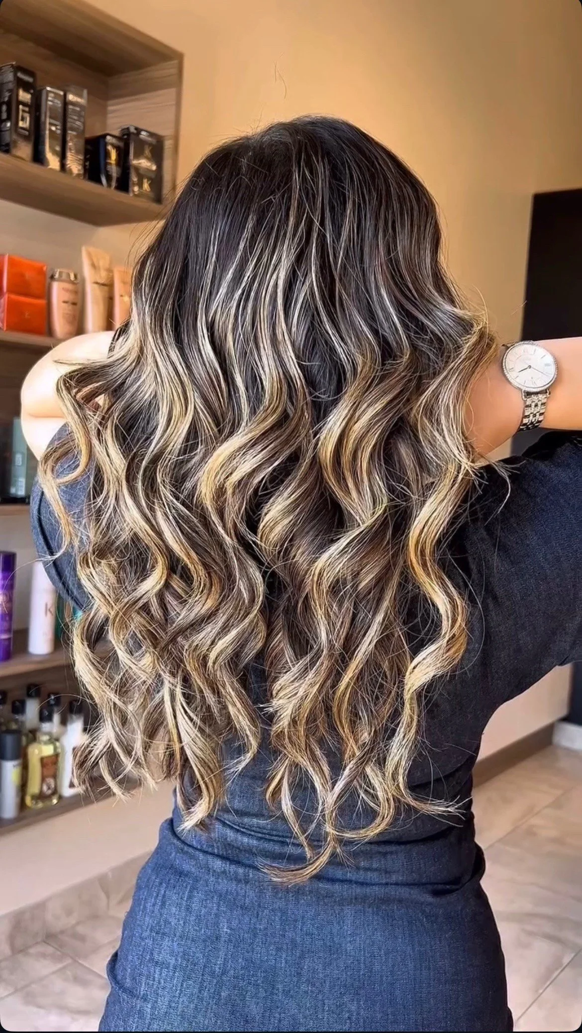Woman with long, wavy hair featuring blonde highlights and darker roots, standing with her back to the camera in a room with shelves of hair products.