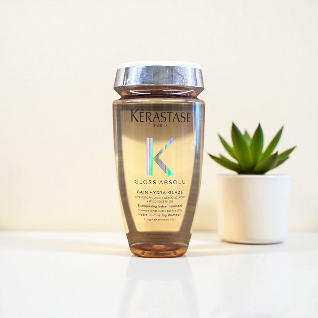 A bottle of Kerastase Gloss Absolu Bain Hydra-Glaze shampoo placed on a white surface next to a small potted succulent plant.