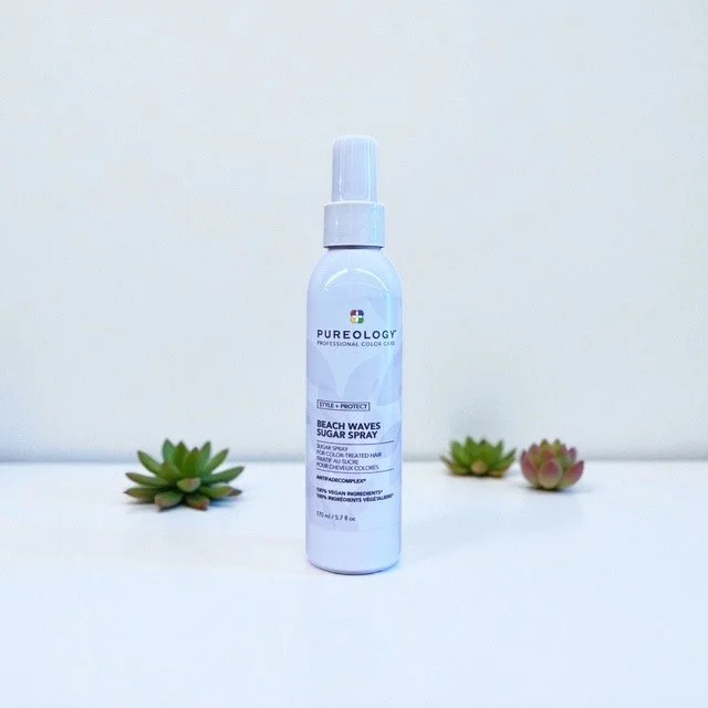 A white bottle of Pureology Beach Waves Sugar Spray on a white surface with small green and pink succulents on either side.