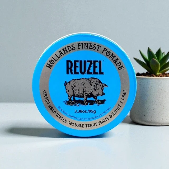 A can of Reuzel Strong Hold Water-Based Pomade with a blue lid, placed on a white surface next to a small potted succulent plant.