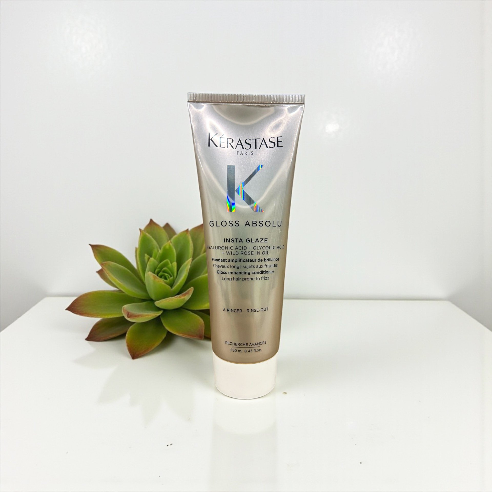 A tube of Kérastase Gloss Absolu Insta Glaze hair treatment standing on a white surface with a green succulent plant behind it.