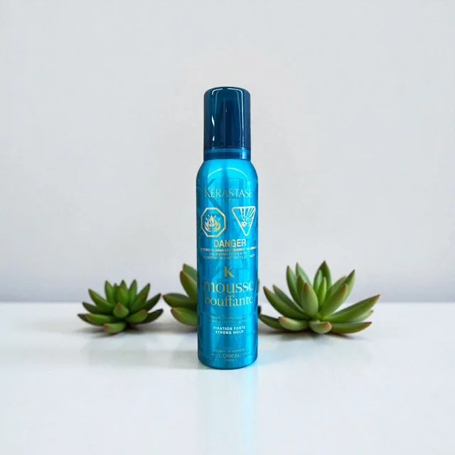 Blue bottle of Kerastase mousse hair product with warning labels, surrounded by green succulent plants on a white surface.