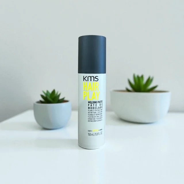 A white canister of KMS Hair Play molding paste with a black cap, placed on a white surface, flanked by two small white pots with green succulent plants in the background.