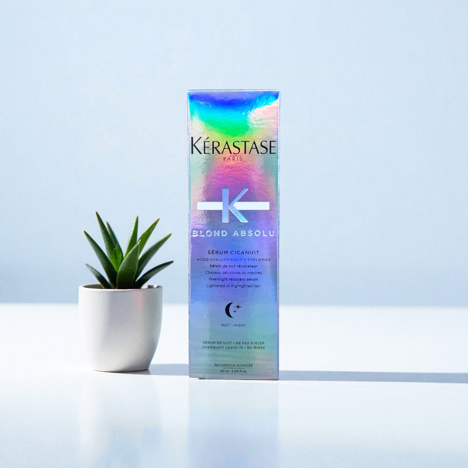A colorful, holographic box of Kérastase Paris Blond Absolu Sérum Cicanuit, standing on a white surface next to a small potted green succulent plant with spiky leaves.