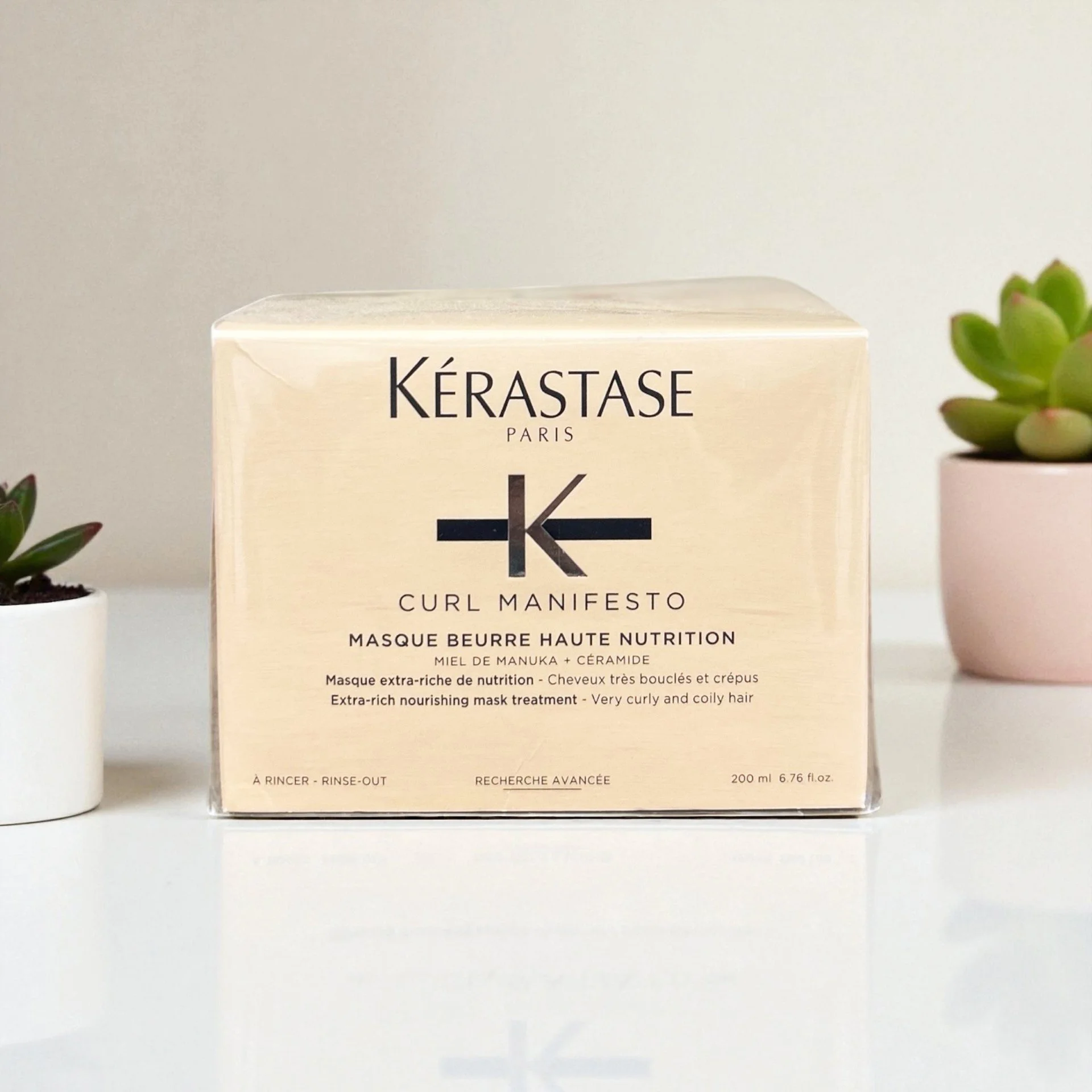 A beige box of Kérastase Curl Manifesto hair mask placed on a white surface with two potted plants on either side.