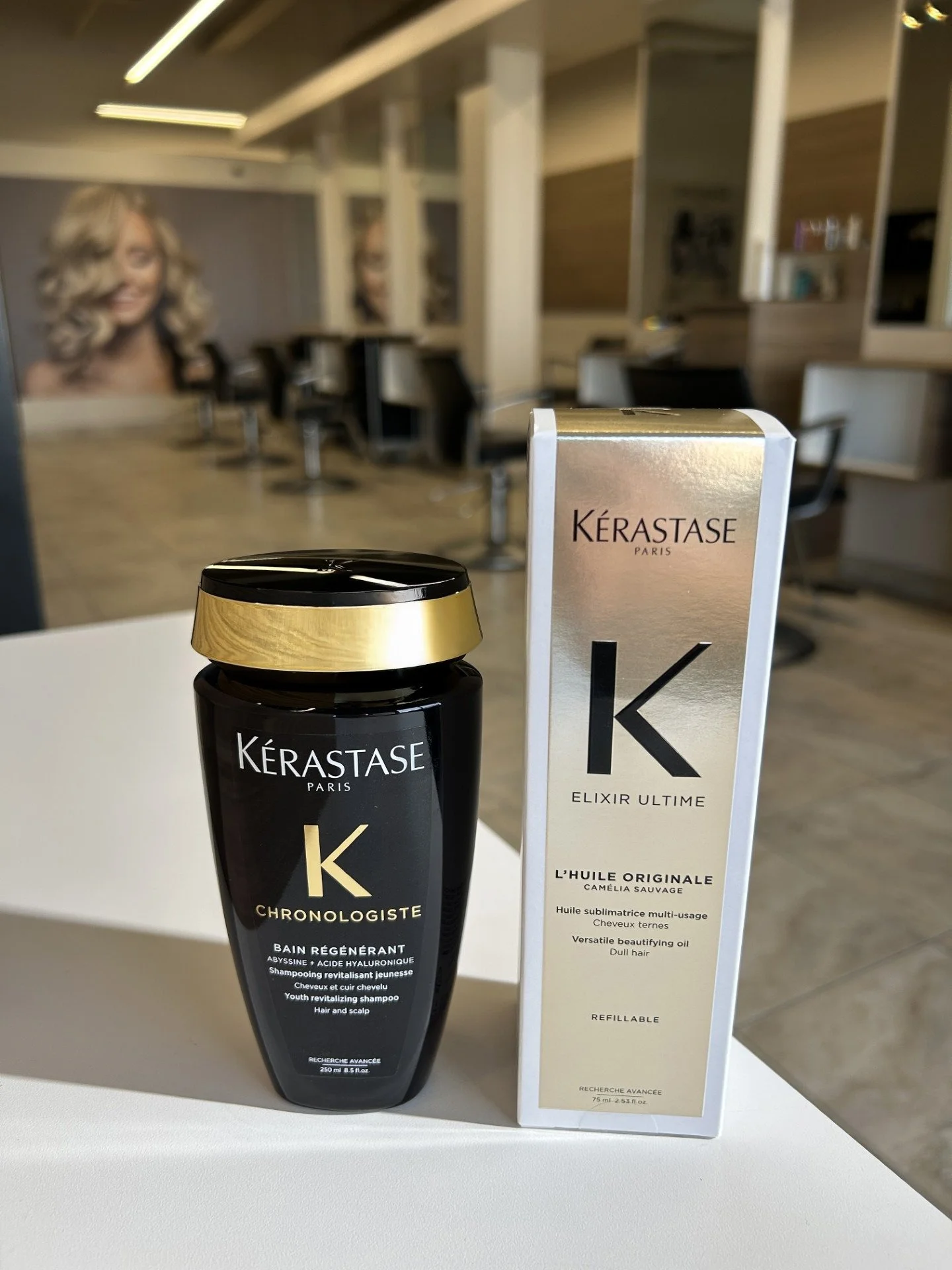 Kérastase hair care products, including Chronologiste revitalizing shampoo and Elixir Ultime beautifying oil, placed on a white surface inside a salon with hair styling chairs and posters of women with styled hair in the background.