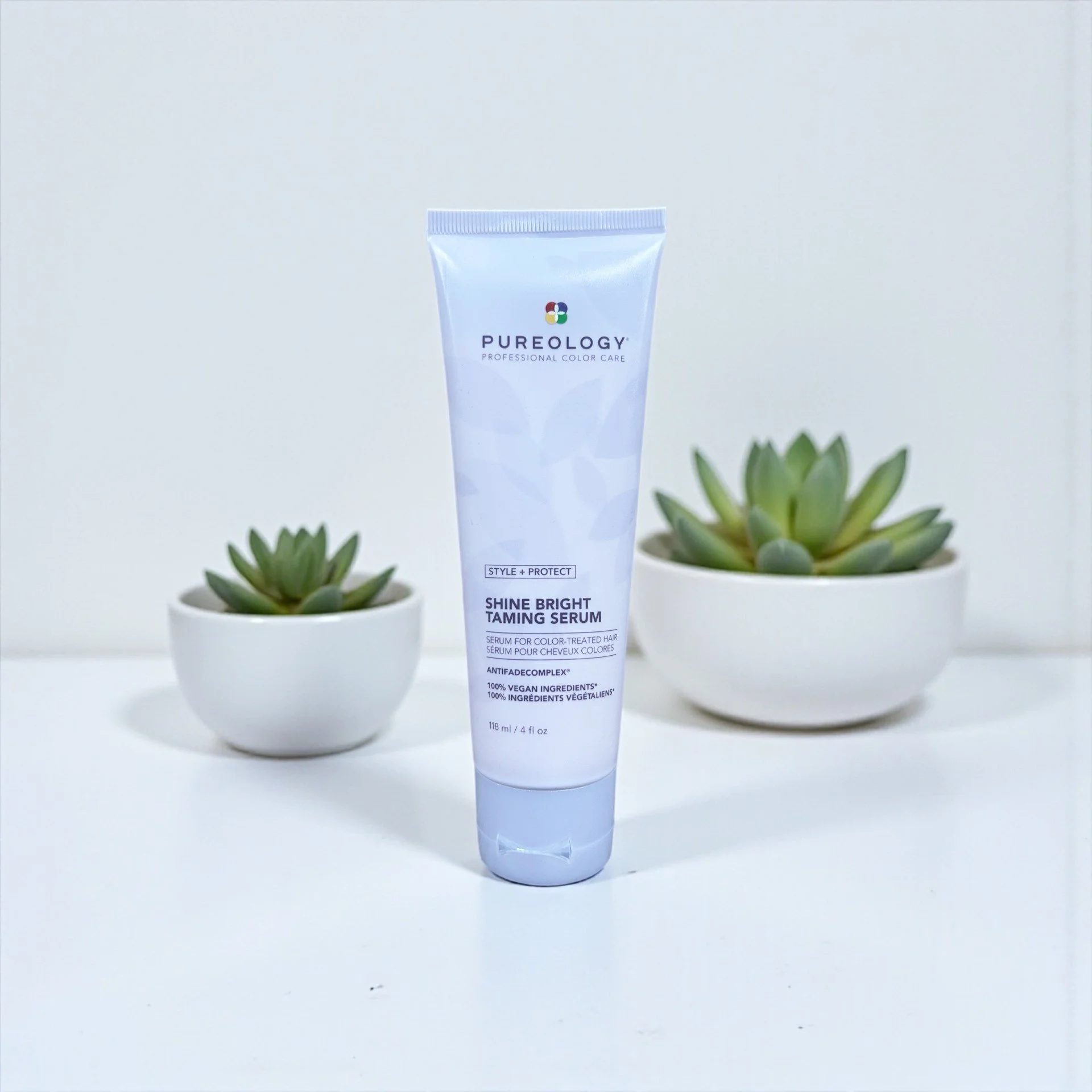 A tube of Pureology Shine Bright Taming Serum standing between two small potted succulents on a white surface against a plain background.