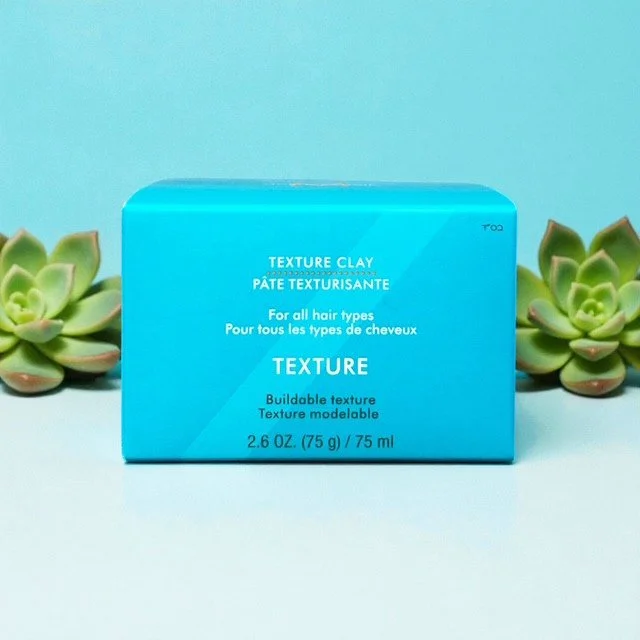 Blue box of Moroccanoil textured clay hair styling product with succulents on each side, light blue background.