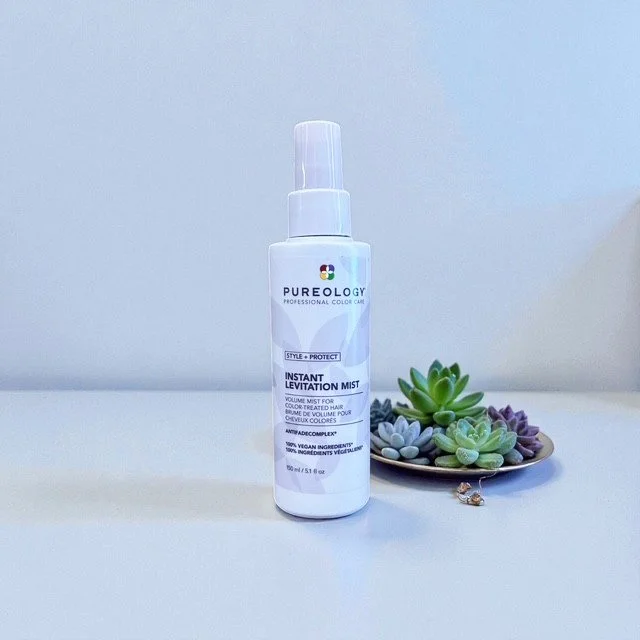 A bottle of Pureology Instant Levitation Mist hair spray next to a small tray of assorted succulents on a white surface.