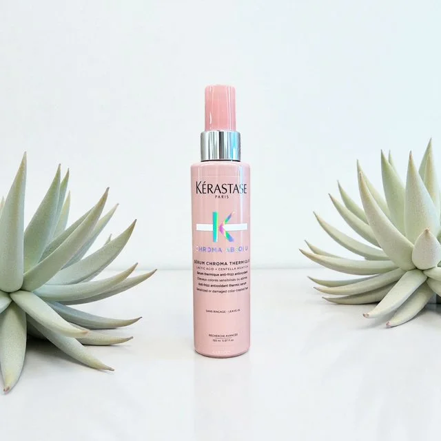 A pink bottle of Kérastase Chroma Absolu Serum Chroma Thermique hair care product is centered, flanked by two pale green succulent plants on a white background.