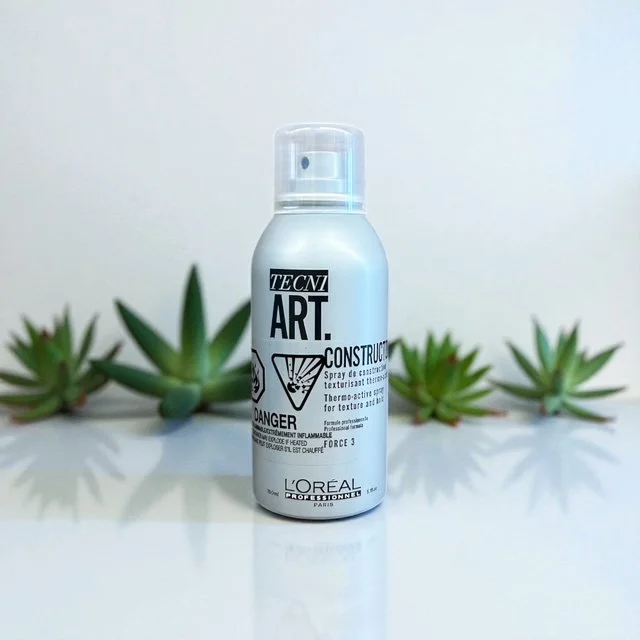 A white bottle of Tecni Art texture spray by L'Oreal Professional on a white surface with green succulents in the background.