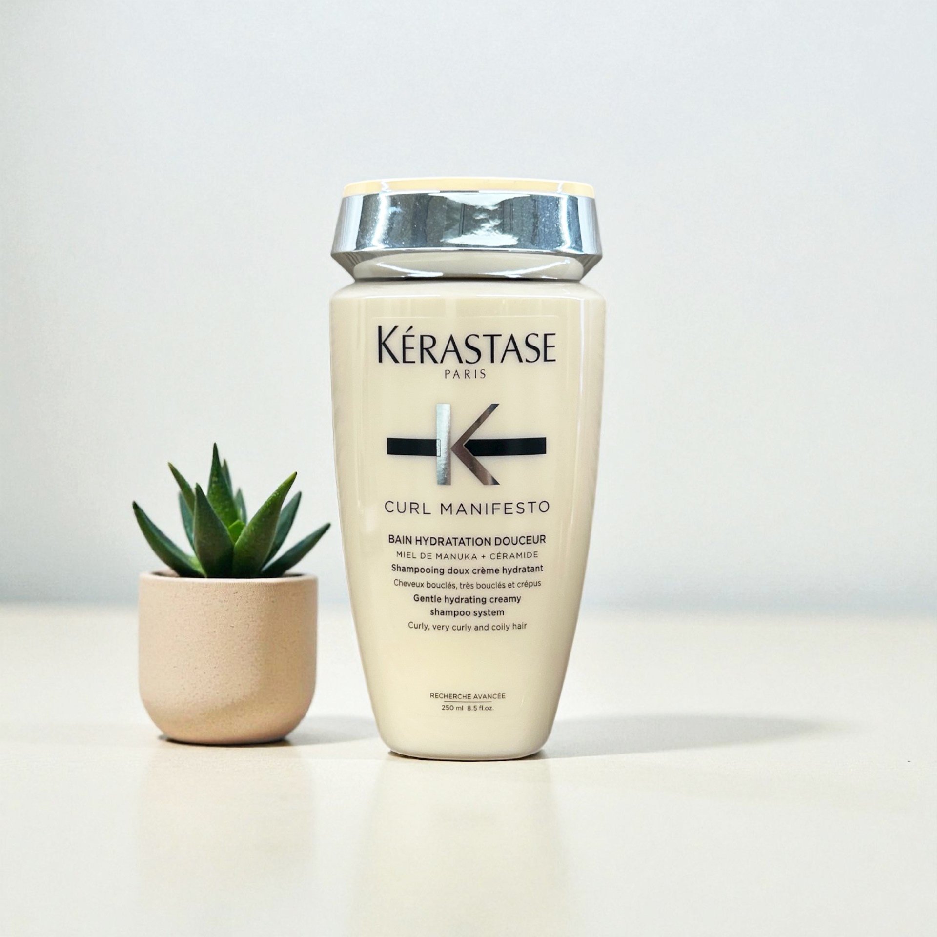 A bottle of Kérastase Curl Manifesto gentle hydrating creamy shampoo next to a small potted succulent plant on a white surface against a plain white background.