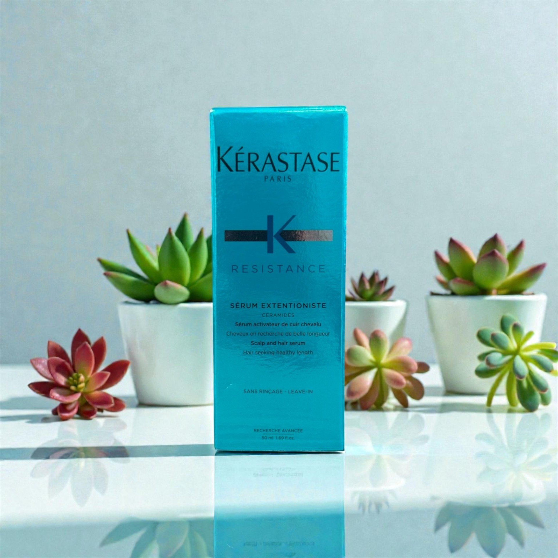 A blue box of Kérastase Resistance hair serum stands on a reflective white surface surrounded by potted succulents in white pots.