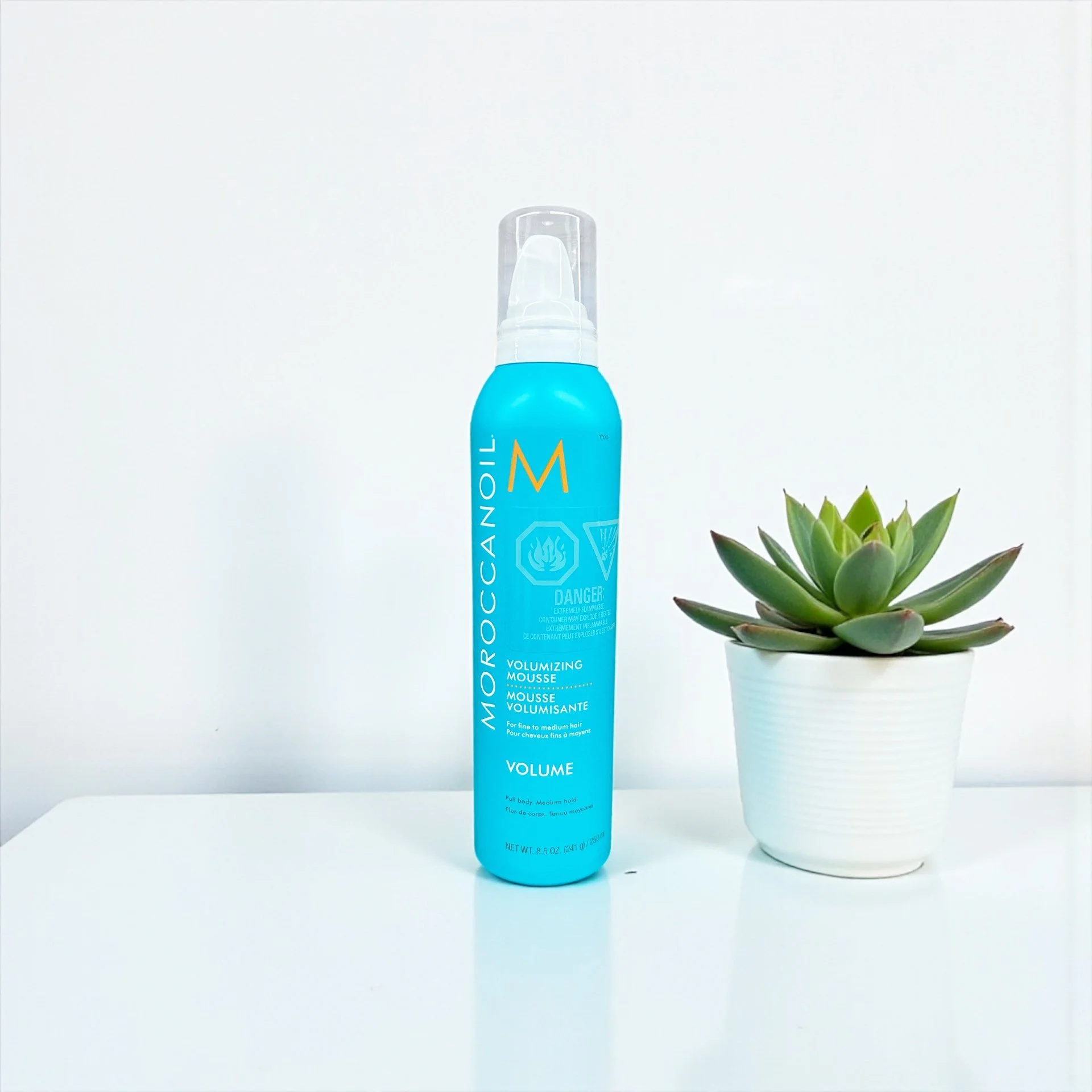 Blue can of Moroccanoil volumizing mousse next to a potted succulent plant, both placed on a white surface with a white background.
