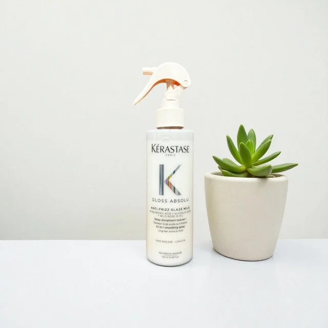 A white spray bottle of Kérastase Gloss Absolu hair product next to a small potted succulent plant on a white surface with a plain white background.