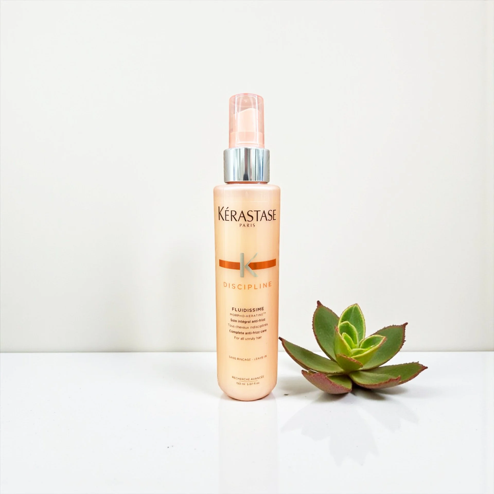 A bottle of Kérastase Discipline Fluidissime anti-frizz hair care spray next to a green succulent plant on a white surface and background.