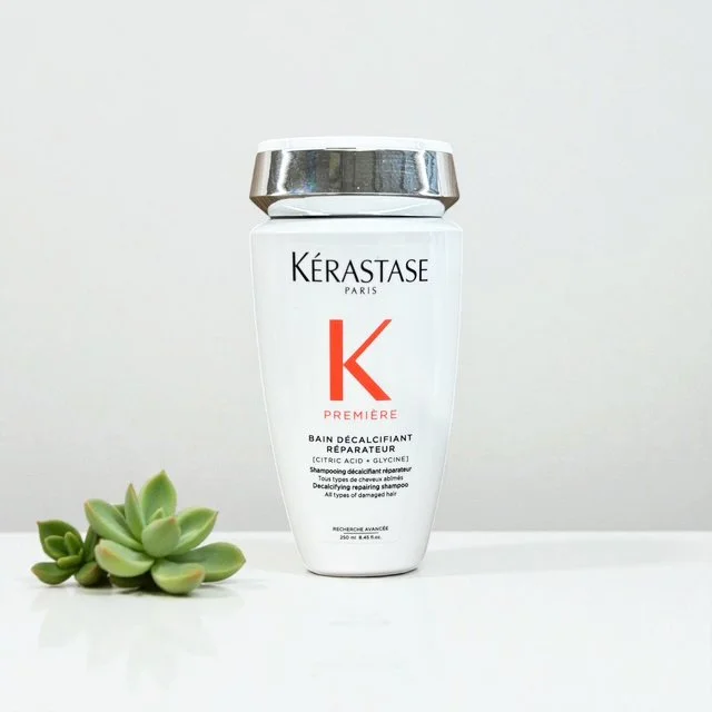 A bottle of Kérastase Paris Genesis Bain Nutri-Fortifiant repairing shower shampoo with a silver cap, placed on a white surface next to a small green succulent plant.