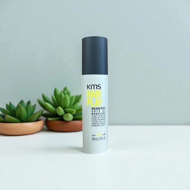 A white and black bottle of KMS Hair Play Molding Paste placed on a white surface with two small green succulent plants in terracotta pots in the background.