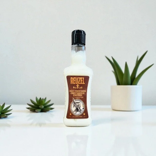 A bottle of Reuzel Daily Conditioner on a white surface with small green succulent plants and a larger potted plant in the background.