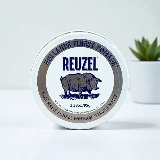 Reuzel clay matte pomade tin on a white surface with a small green succulent plant in the background.