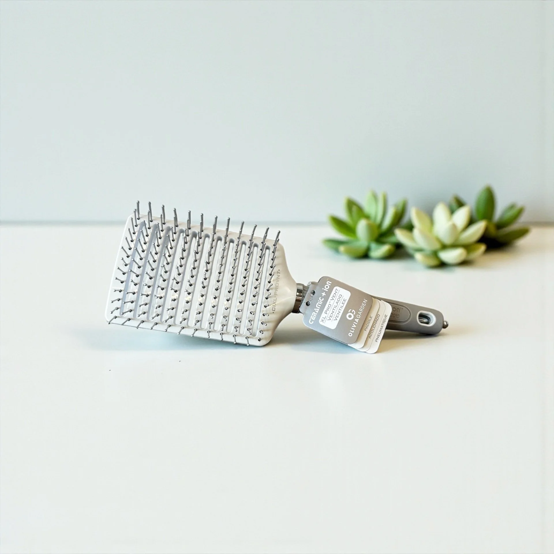 A metal hairbrush with a handle lying on a white surface, with a small tag attached, and a green succulent plant in the background.