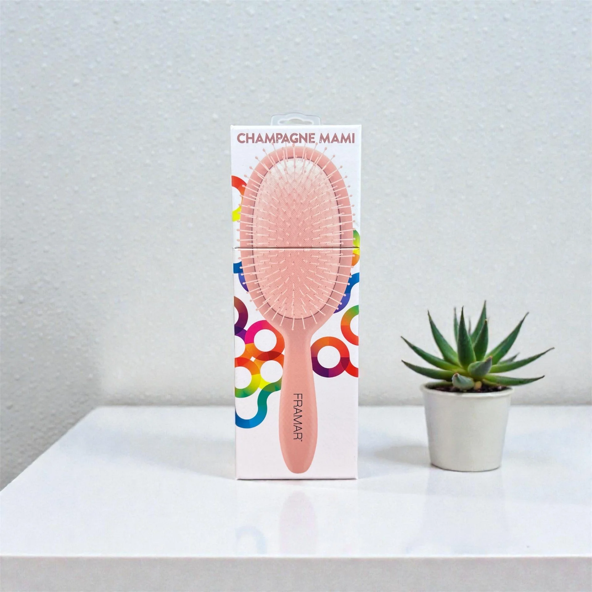 A pink hairbrush in front of colorful circular designs, next to a potted succulent plant on a white surface with a textured gray wall background.