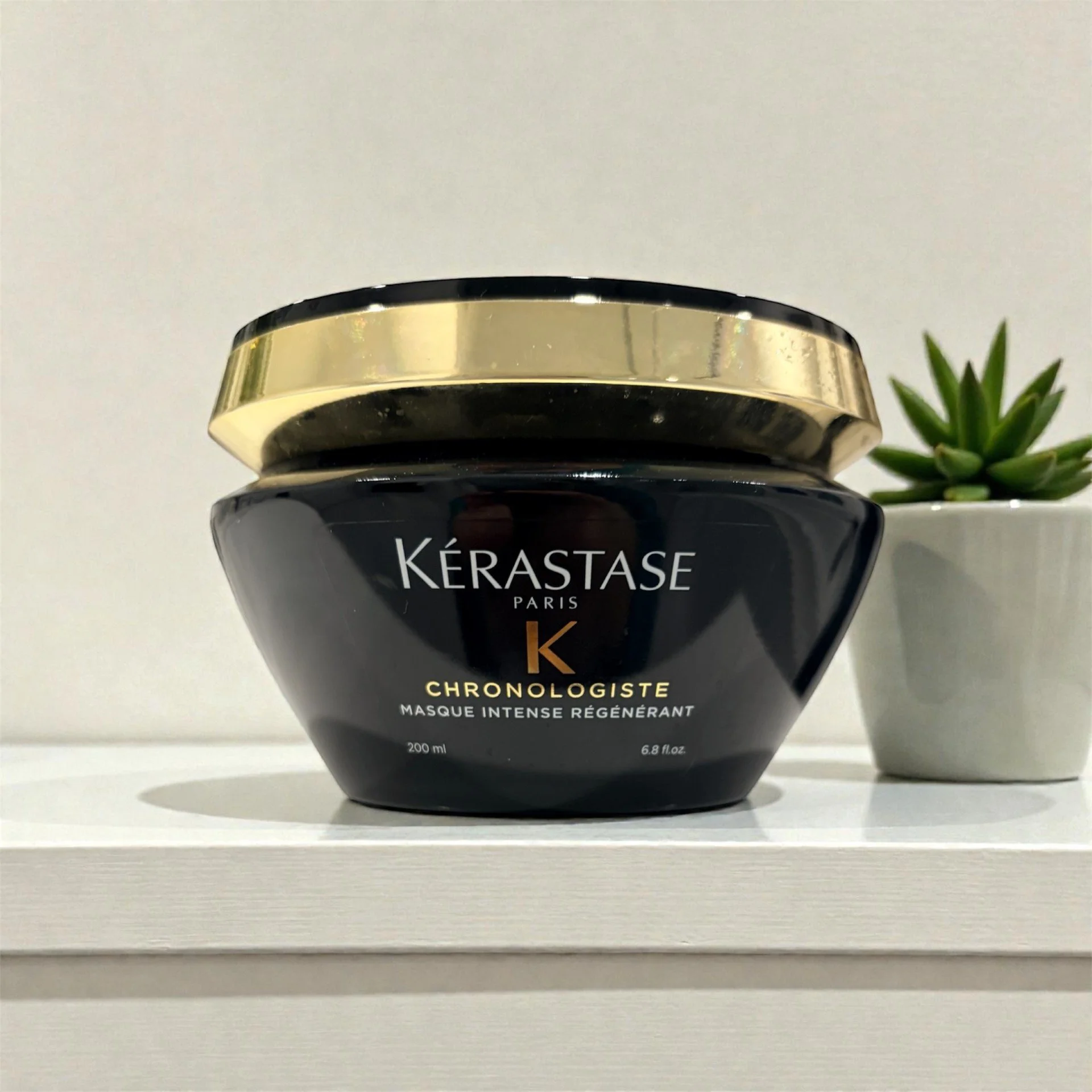 A black and gold jar of Kerastase Paris Chronologiste mask placed on a white shelf next to a small potted succulent plant.