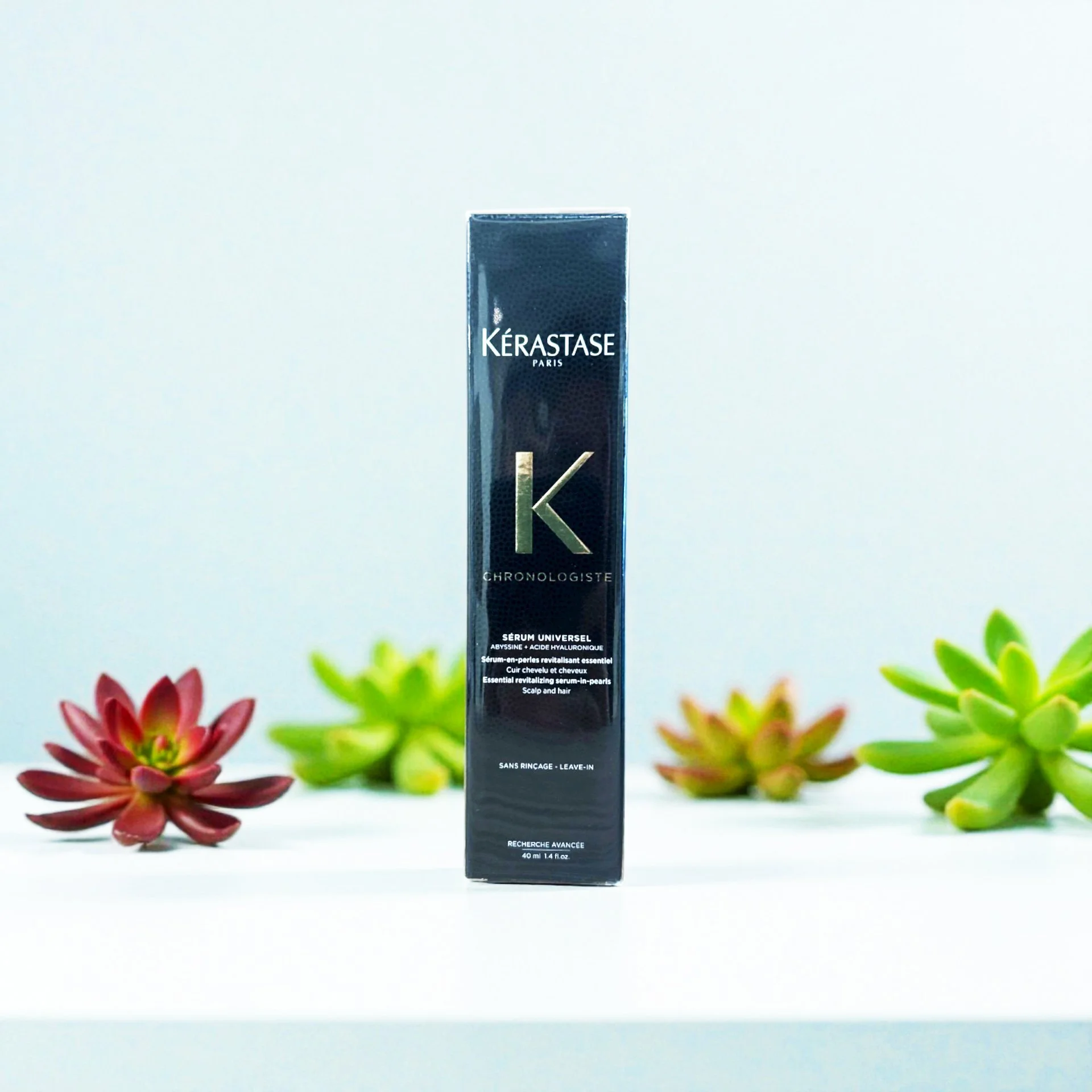 A tall black box of Kerastase Chronologiste serum stands on a white surface, with pink and green succulent plants in the blurred background.