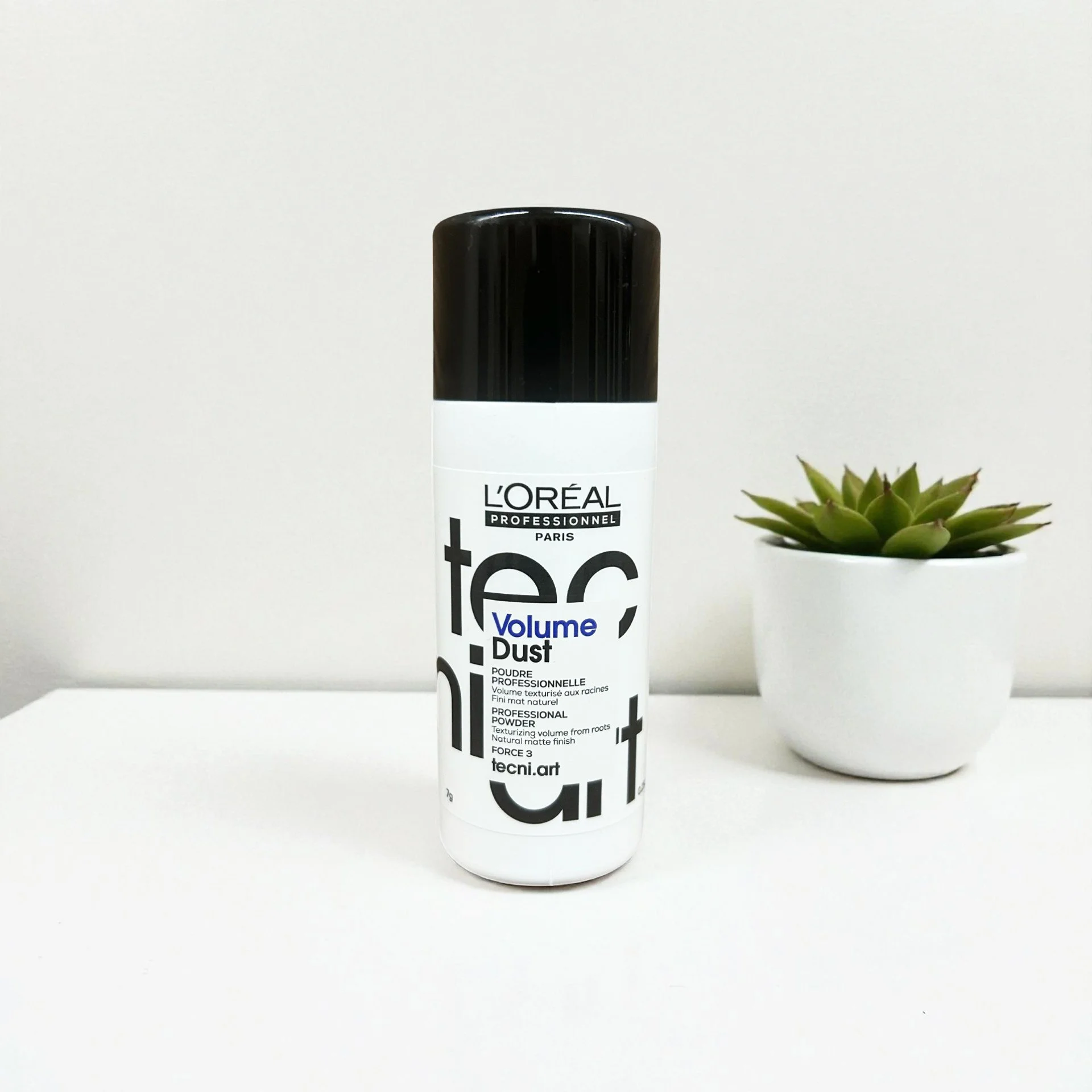 Can of L'Oreal professionnel volume dust spray next to a small potted succulent on a white surface with a white background.