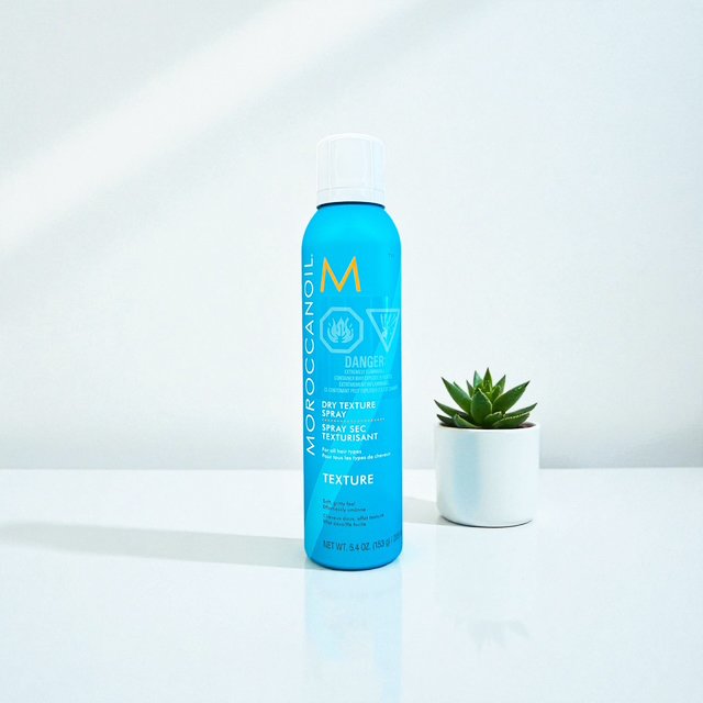 Blue spray bottle labeled Moroccanoil Dry Texture Spray with a white cap and a small potted succulent plant beside it on a white surface.