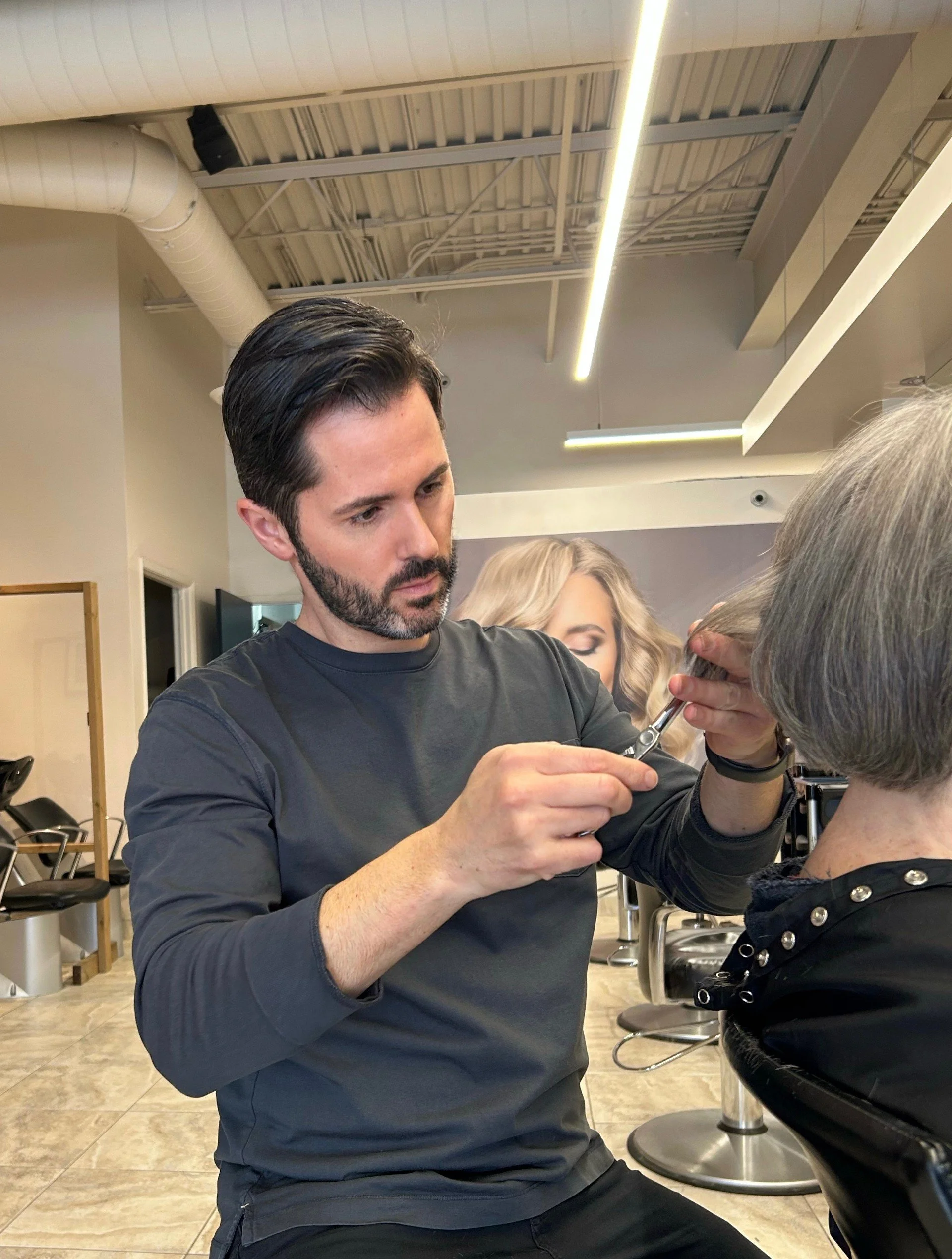 A male hairstylist with dark hair and a beard cutting an older woman's hair in a salon.