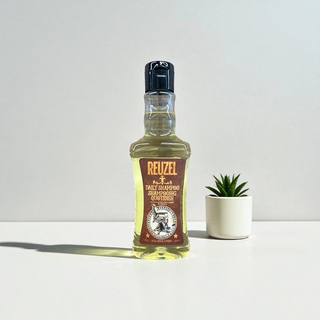 A bottle of Reuzel daily shampoo soap with a black cap and a yellow liquid inside, positioned on a white surface next to a small potted succulent plant.